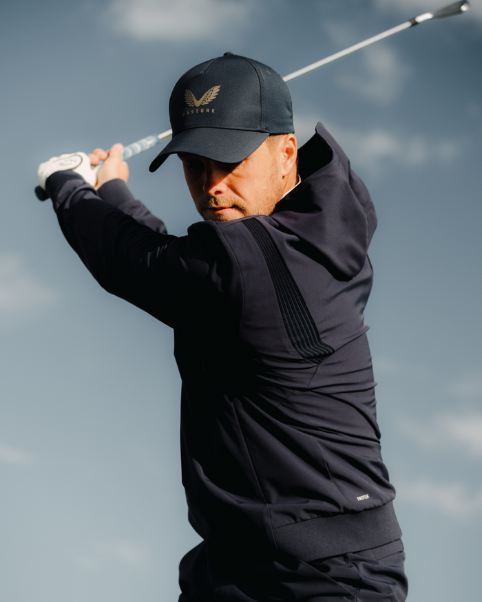 Golfer in midnight navy performance hoodie swinging a golf club against a blue sky
