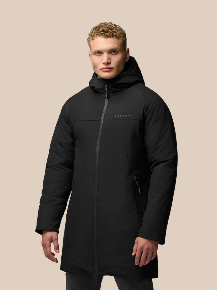 Model wearing black longline hooded jacket with front zip and zip pockets, featuring ProTek water resistance and Castore branding on chest and hood.