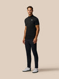 Full-body shot of a man in a black Pinnacle Pique Stripe Polo with all-over stripes, dark trousers and white golf shoes.