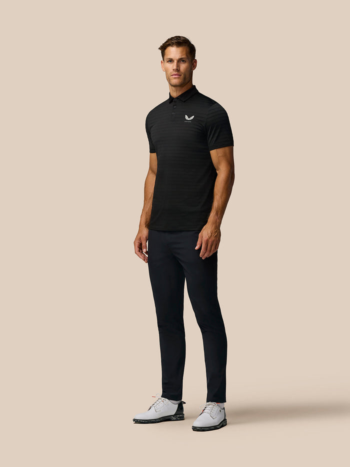 Full-body shot of a man in a black Pinnacle Pique Stripe Polo with all-over stripes, dark trousers and white golf shoes.