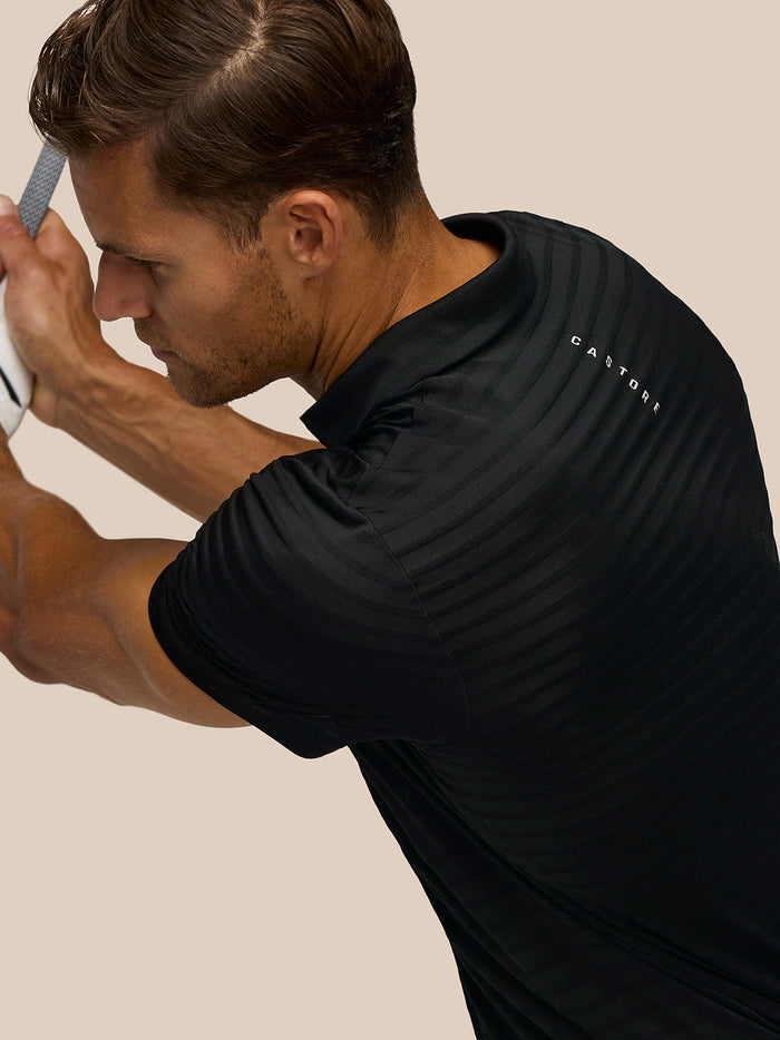 Close-up of a golfer in a black striped polo gripping a club with Castore branding visible on the back.