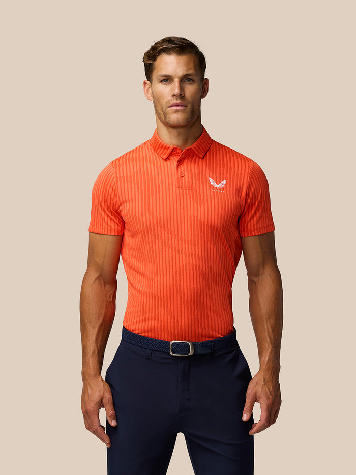 Front view of a man wearing a bright orange striped golf polo with Castore logo on left chest, tucked into navy trousers