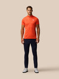 Male model wearing Golf Pinnacle Glow Polo Bright Orange with striped knit and Castore logo on left chest, navy trousers.