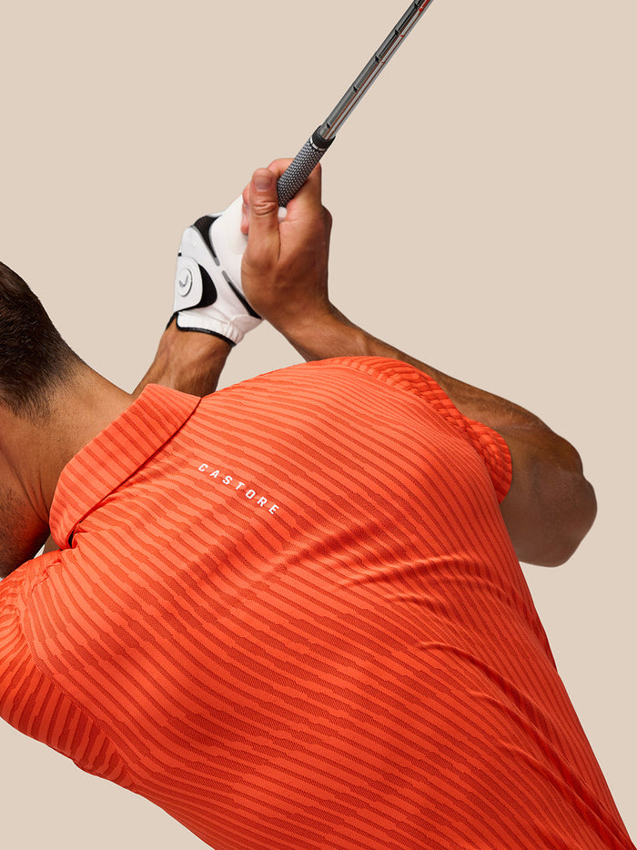 Rear view of a golfer wearing a bright orange striped Pinnacle Glow Polo while gripping a golf club