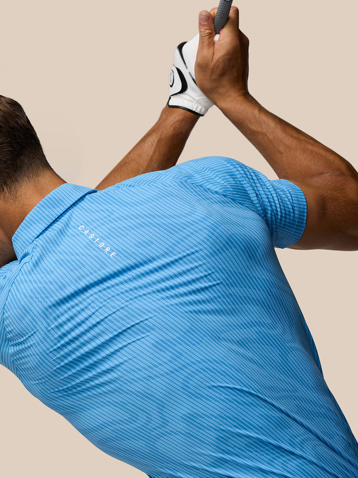 Male golfer wearing Cool Blue Golf Club Glow Printed Polo seen from behind mid swing with Castore logo on the chest.